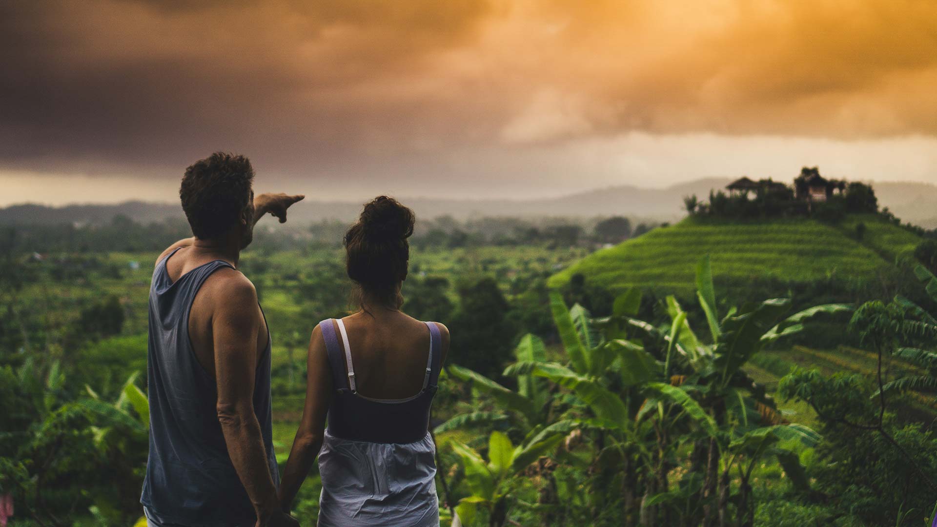 Ein Mann und eine Frau stehen auf Bali im Dschungel auf einem Berg und zeigen auf eine tolle Villa am anderen Ende der Schlucht